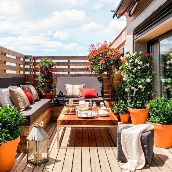A summer balcony with a wooden deck and furniture, potted blooms and greenery and bright pillows plus candle lanterns.