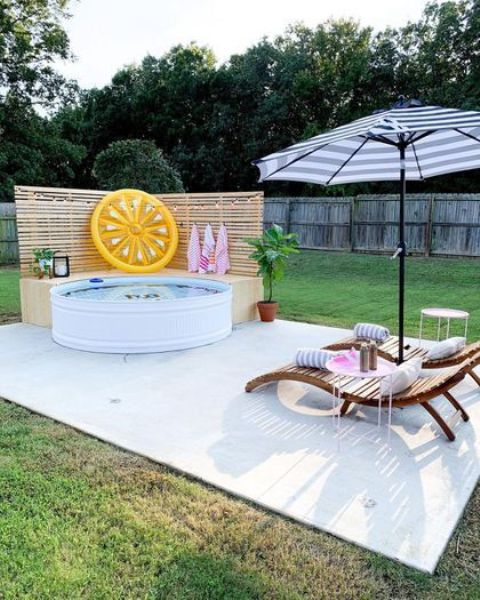A tiled deck with a stock tank pool, a wooden bench, greenery and a float, loungers and an umbrella.