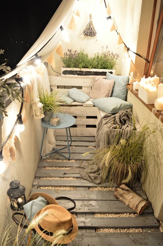 A tiny cozy balcony with a wooden floor, pallet furniture and pots, potted greenery and blooms and lights and candles.