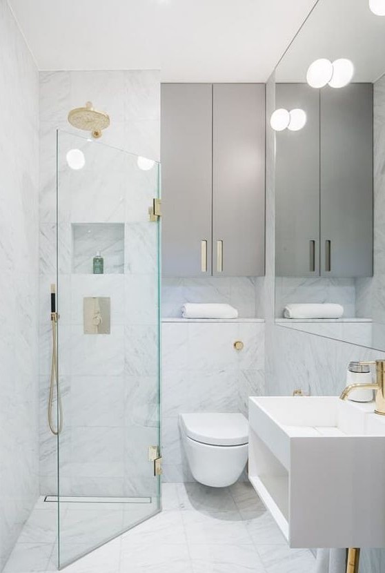 A tiny minimalist bathroom with white marble tiles, sleek grey cabinets, gilded touches and a triangle shower space.
