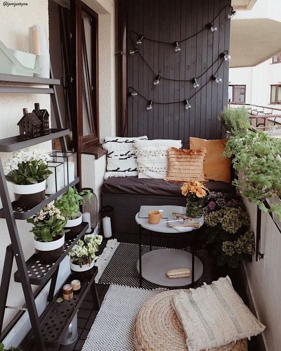 A tiny monochromatic balcony with black and white walls, blakc furniture, potted greenery and blooms and various pillows and rugs.