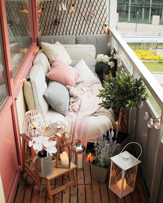 A tiny narrow balcony with pastel textiles and pillows, potted greenery and blooms, candle lanterns and wooden furniture.