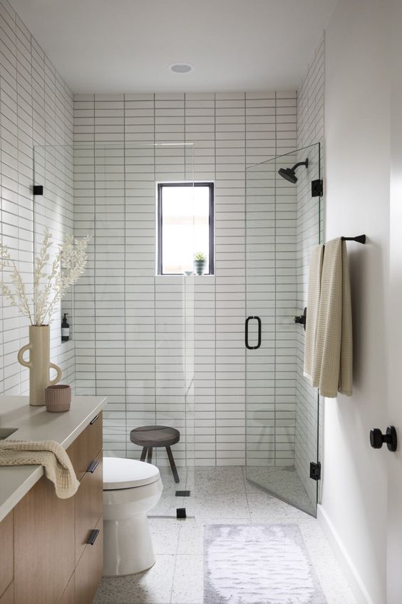 A tiny neutral bathroom with white skinny tiles and a shower space, a stained vanity and black fixtures plus a small window.