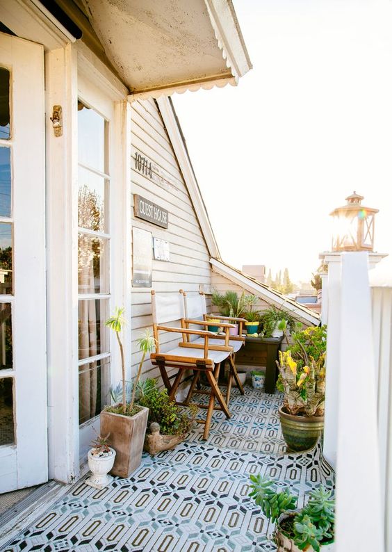 A tiny welcoming balcony with a bright rug, folding chairs, potted greenery and blooms to enjoy some coffee.