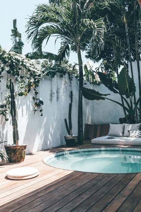 A tropical outdoor space with a wooden deck, a daybed with pillows, a stock tank pool and some potted plants and trees.