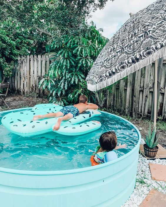 A turquoise stock tank pool with a float, an umbrella, some greenery and potted plants is a very cool space.