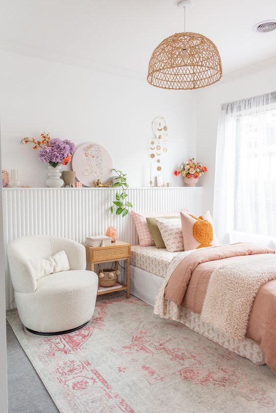 A vivacious teen girl bedroom in neutrals with a paneled shelf with blooms and art, a bed with printed bedding, a white chair and a woven pendant lamp.