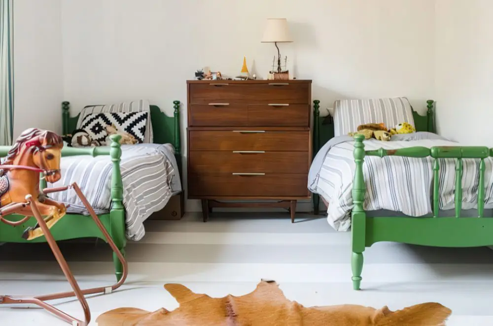 A charming shared boys' room featuring twin green wooden beds with spindle details, striped bedding, and a mid-century modern dresser. The space exudes a nostalgic feel, enhanced by a wooden rocking horse and playful plush animals.
