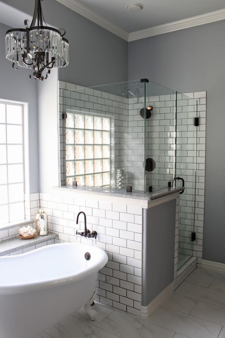 A grey bathroom with white subway tiles, shower space with a half wall, a crystal chandelier and a vintage bathtub.