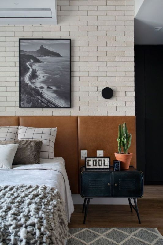 This modern space is done with brick walls, a bed with an extended brown leather headboard, a metal cabinet nigthstand and some decor.