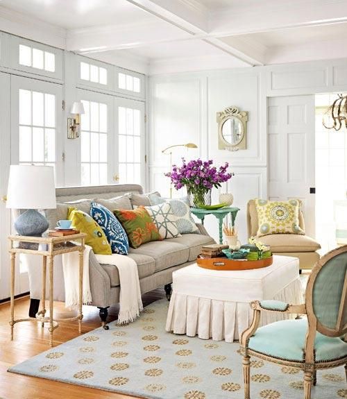A welcoming neutral living room done with bright and colorful pillows, with a printed rug and some blooms in a vase.