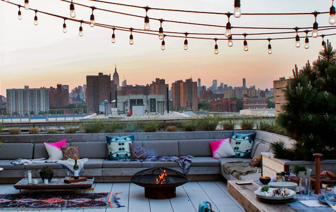 A rooftop summer terrace with a large built-in bench, tables and a fire pit, colorful pillows and string lights over the space.