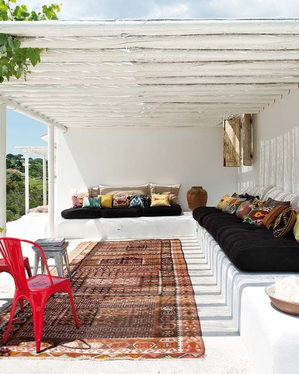 A white summer terrace with a built-in bench, dark upholstery and colorful pillows, a boho rug and a cool sea view.