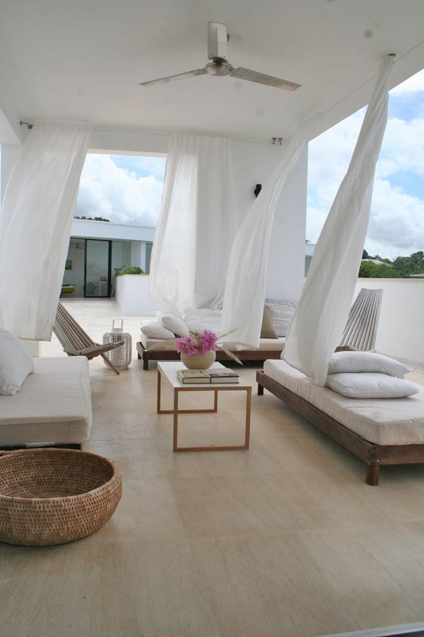 A neutral summer terrace with low wooden furniture, neutral upholstery and pillows, white curtains, candle lanterns and some blooms.