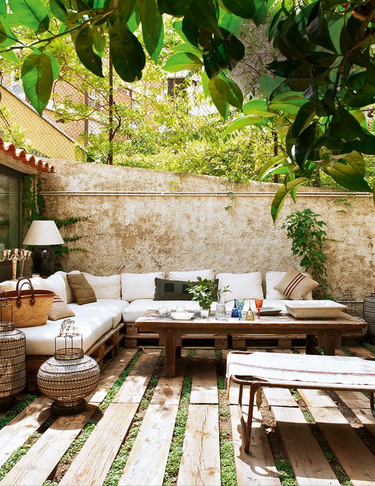 A welcoming Mediterranean summer terrace with pallet and rattan furniture, neutral upholstery and textiles, candle lanterns and greenery.