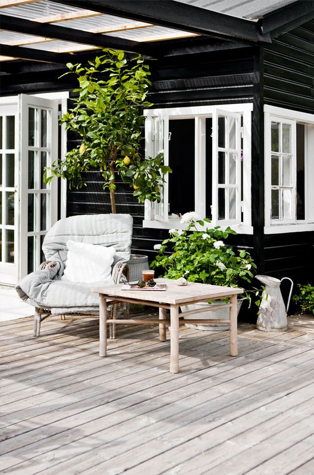 A summer Nordic terrace with rattan furniture, cozy neutral upholstery, potted greenery and a large tree.
