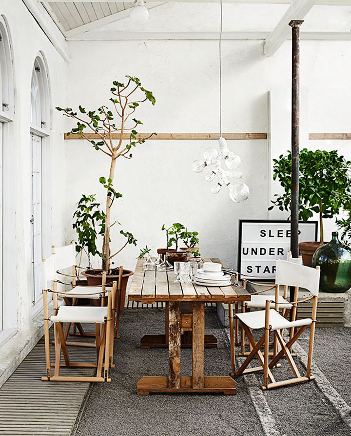 A Nordic summer terrace in neutrals, with wooden and rattan furniture, potted greenery and trees and a bulb chandelier.