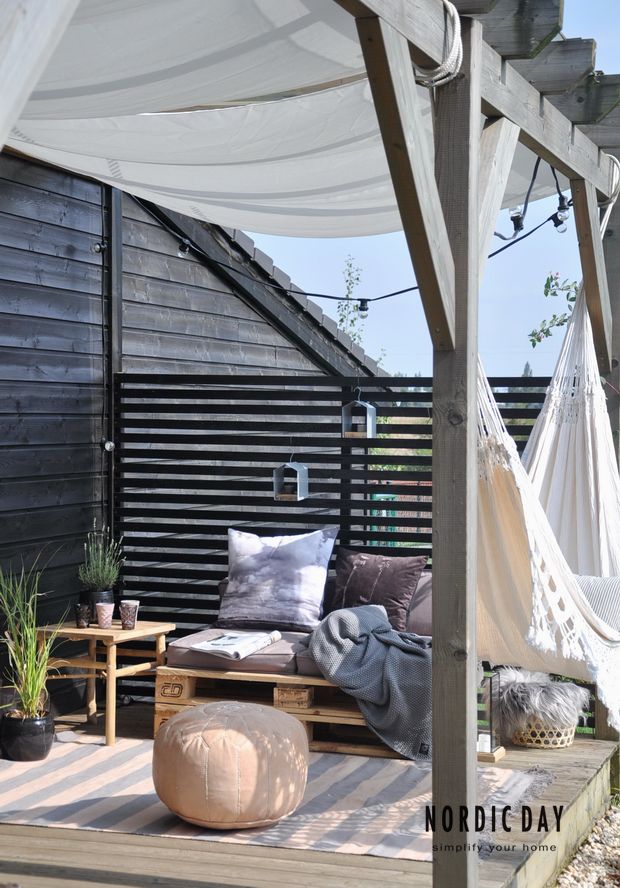 A small Nordic summer terrace with dark walls, pallet and wooden furniture, dusty textiles and a hammock plus ottomans.