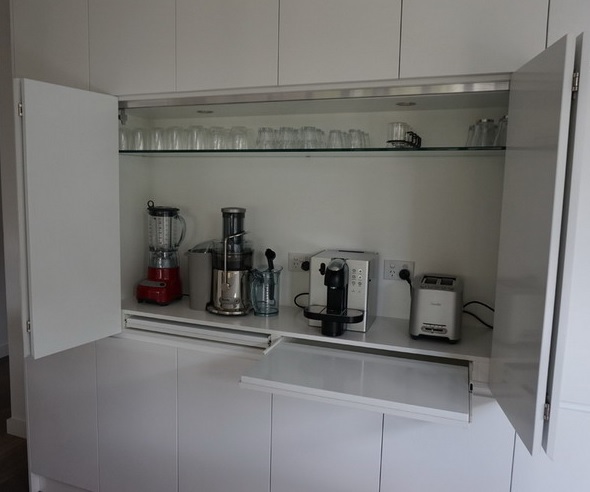 A white cabinet with a glass and retractable shelves and several different appliances is a real cooking and coffee station.