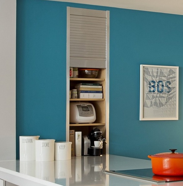 A built-in storage unit that contains various appliances, books and tableware and is closed with a sliding shutter.