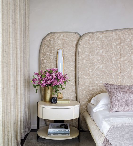 The refined neutral bedroom is done with a boucle bed and a printed panel headboard with lights, a round nightstand and neutral curtains.
