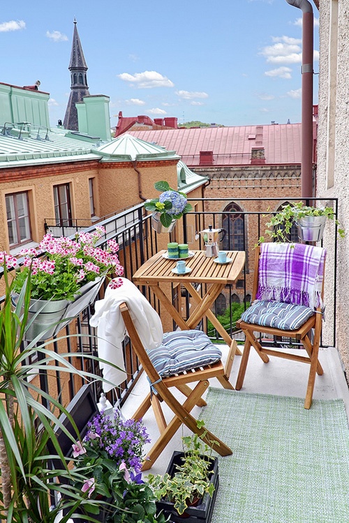 A cheerful summer balcony with bright textiles, potted blooms and greenery and simple wooden folding furniture.