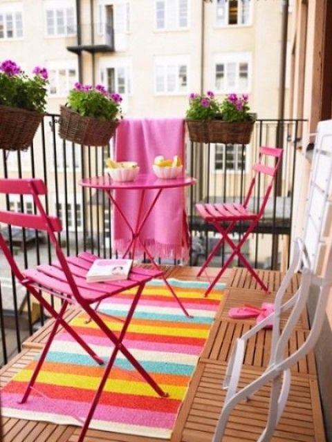 A small and bright summer balcony with a bright rug and blanket, bold pink furniture and potted blooms.