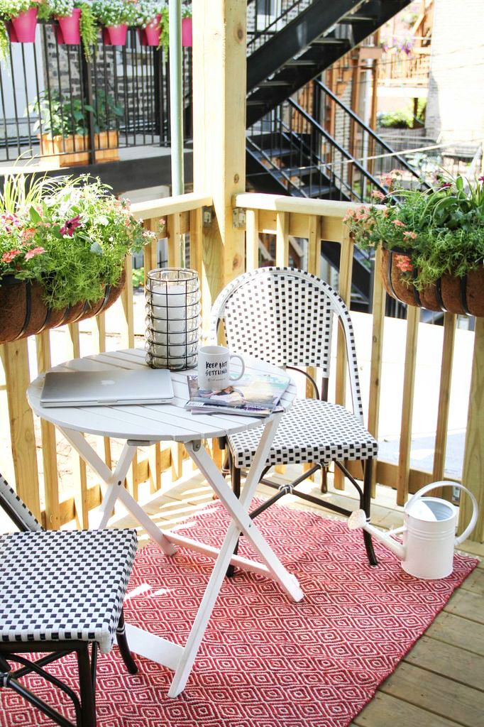 A small and bright summer balcony with dark wooden chairs, a white table, a bright rug and potted greenery and blooms on the railings.