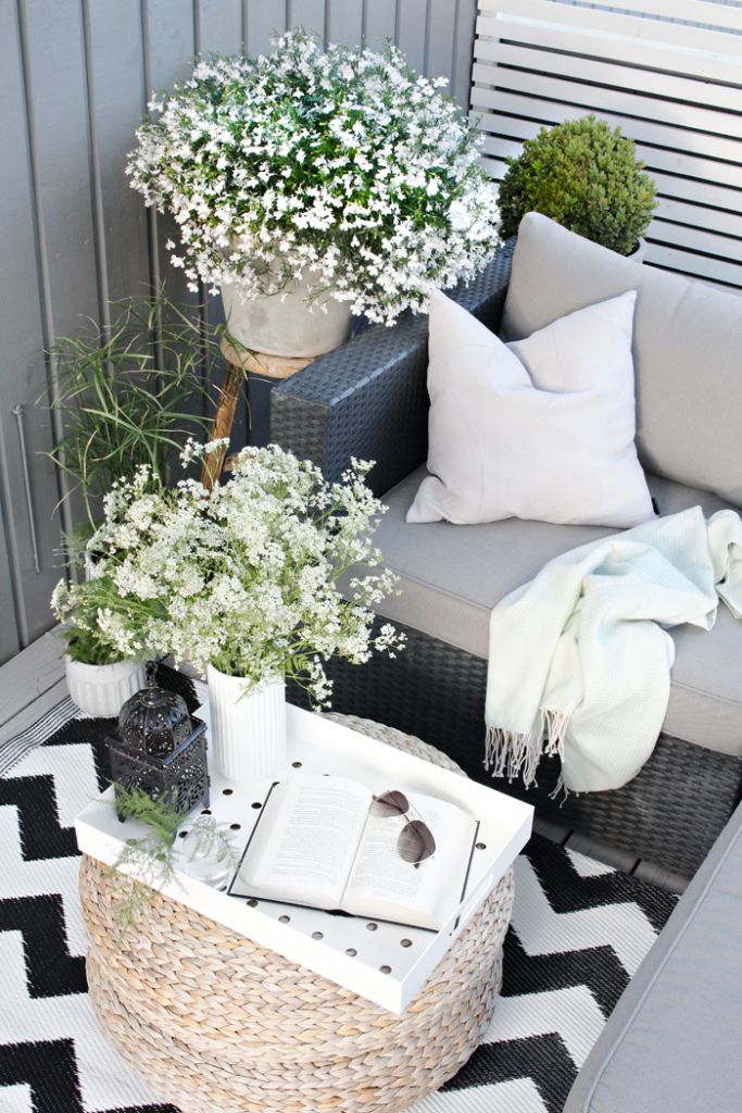 A monochromatic summer balcony with neutral and black wicker furniture, neutral upholstery, potted blooms and greenery and geometric prints.