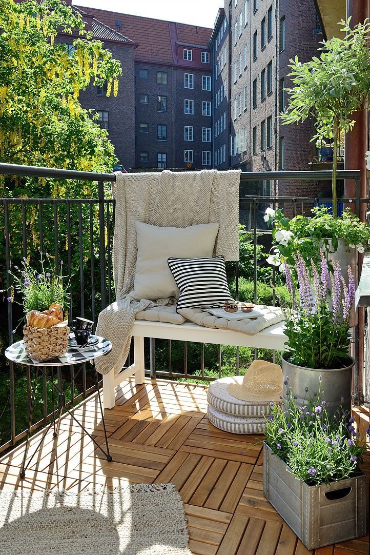 A Nordic summer balcony with a white bench, a metal table, potted greenery and blooms, neutral and printed textiles.