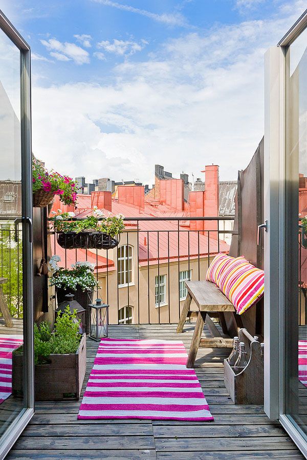 A small yet bright summer balcony with with a simple wooden bench, colorful striped textiles, potted greenery and blooms to catch some sun.