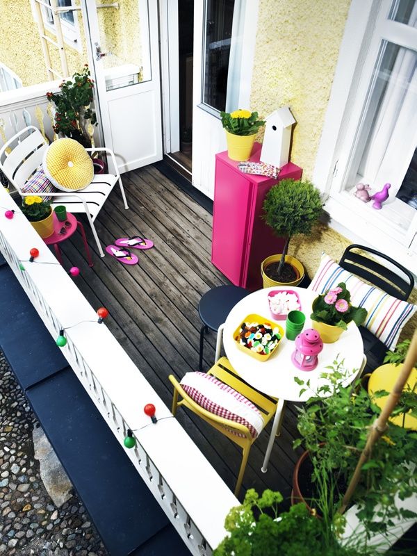 A modern colorful balcony with a wooden floor, metal chairs, colorful textiles and potted greenery.