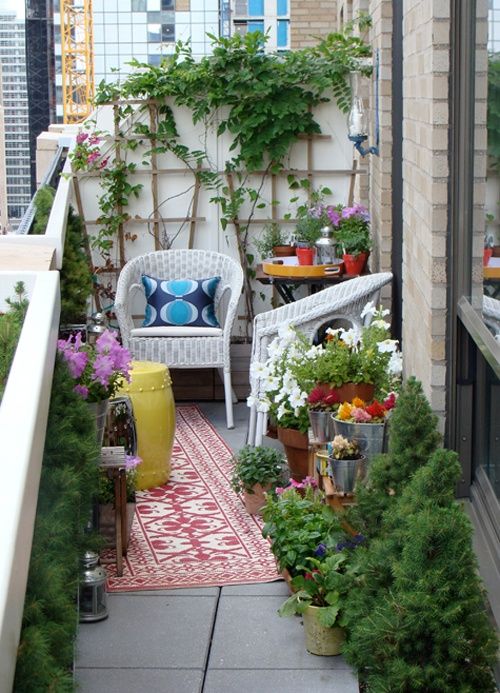 A bright summer balcony with lots of greenery and blooms in pots, with bright rugs and white wicker furniture, bright textiles and accessories.