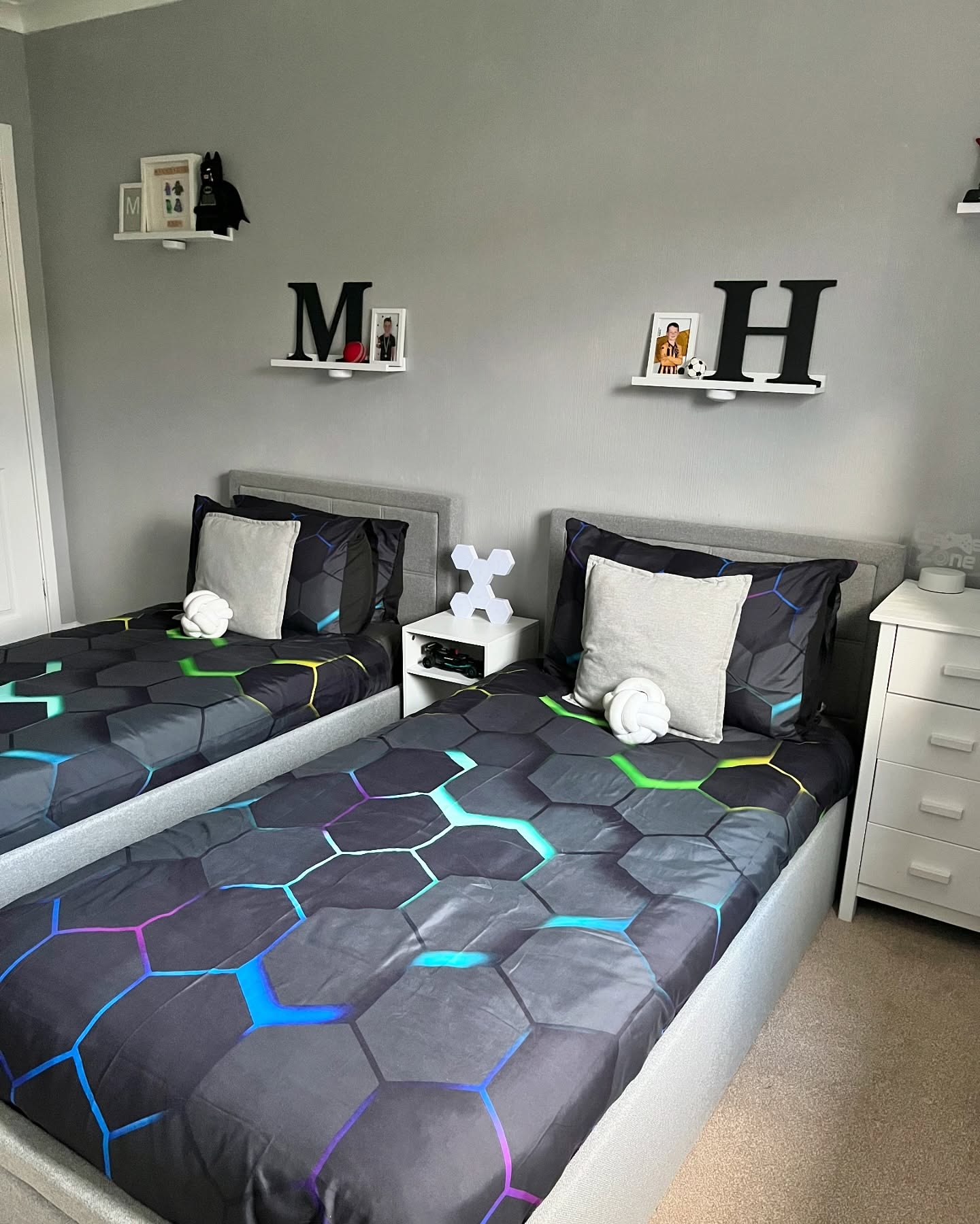 A futuristic gaming-themed bedroom featuring twin beds with neon-patterned bedding. Floating white shelves display superhero figurines, while a dark grey color scheme enhances the modern aesthetic. (@our_house_is_home).
