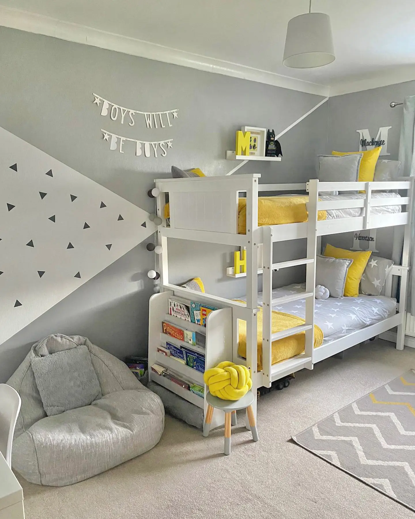 A lively shared bedroom featuring a white bunk bed, geometric wall patterns, and cheerful yellow accents. A cozy bean bag corner and a well-organized bookshelf add a functional touch.
(@our_house_is_home).