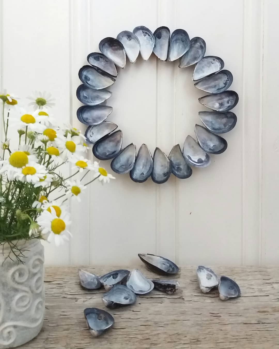 A minimalist wreath crafted from deep blue mussel shells, arranged in a circular pattern that mimics delicate petals, perfect for beach-inspired decor.

(@myplanb.no).