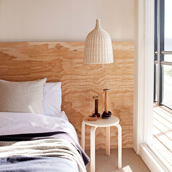 The neutral bedroom shows off a bed with a stained extra wide plywood headboard, a stool as a nightstand and a woven white pendant lamp.