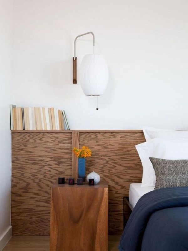 The bedroom is finished off with a stained plywood headboard and a stump nightstand, with books and a large lamp.