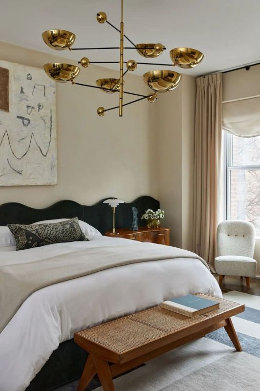 The tan bedroom is done with a dark green wavy headboard, a cane bench, a neutral boucle chair and a gold chandelier.